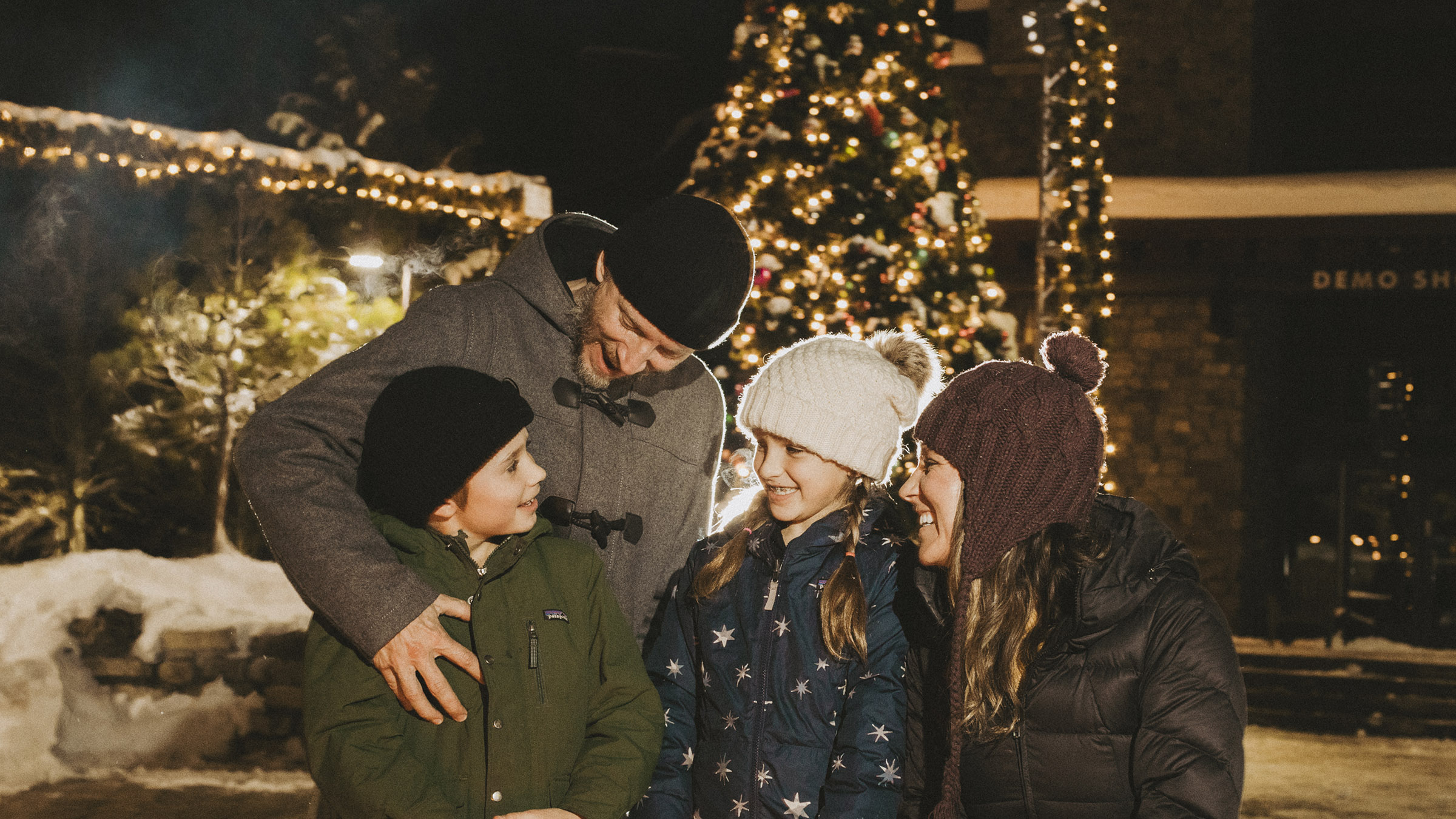 A smiling family poses together near the illuminated holiday tree during the winter tree-lighting event in Mammoth Lakes, California surrounded by festive lights and snowy charm.
